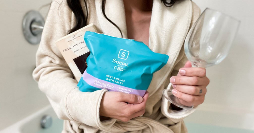 woman holding bath salts, wine glass and book