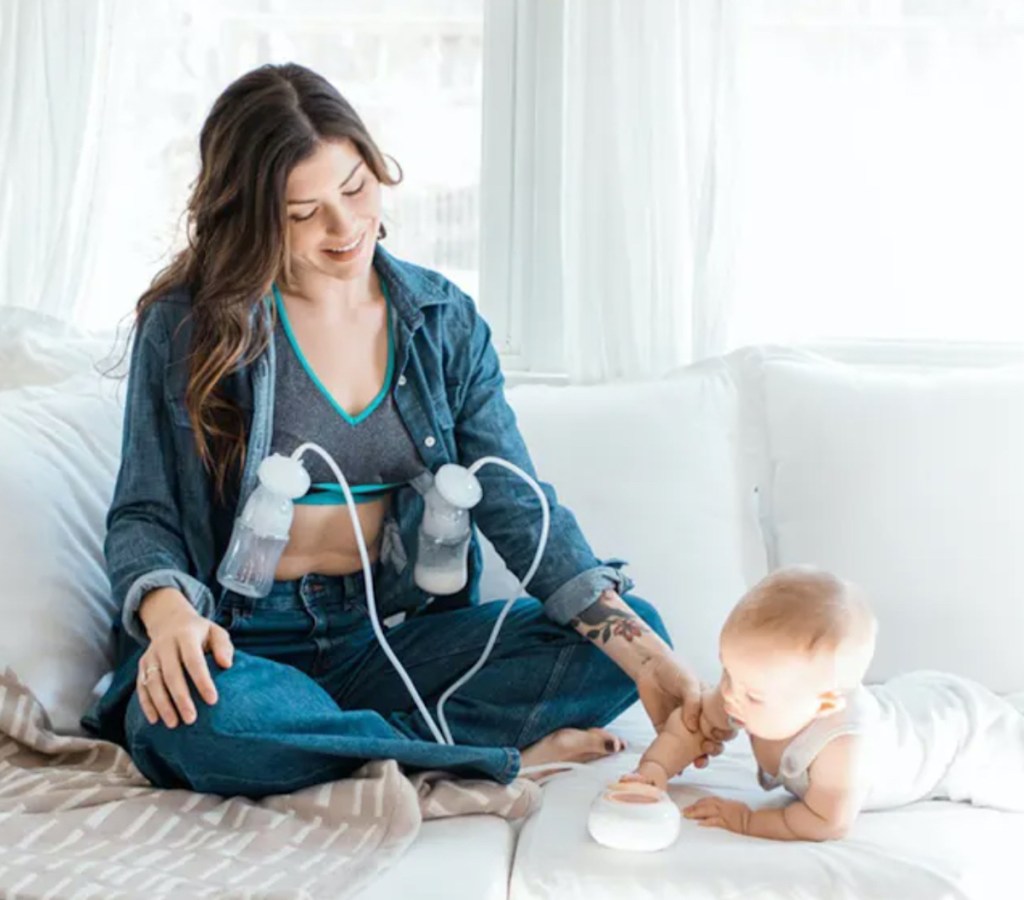 woman with breastpump on chest smiling at baby on couch