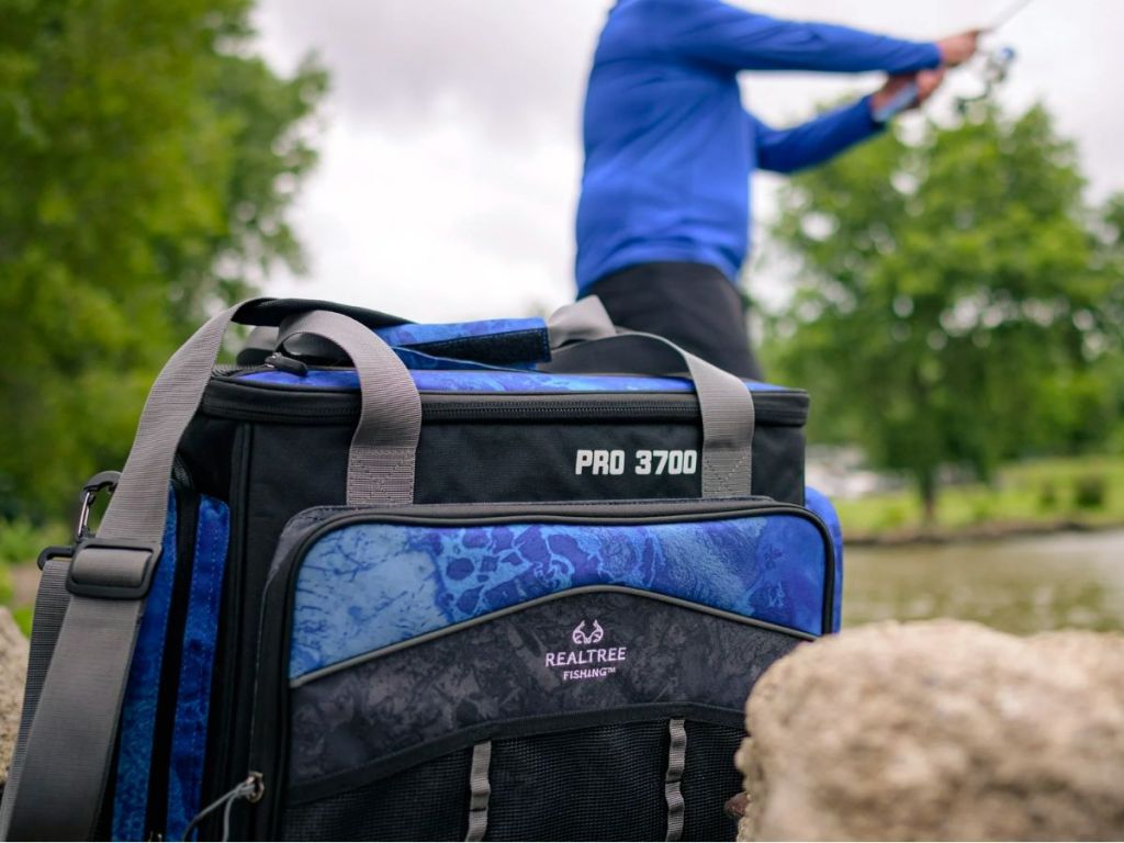 a realtree tackle bag sitting on the bank of a lake with a man in the background fishing with a reel