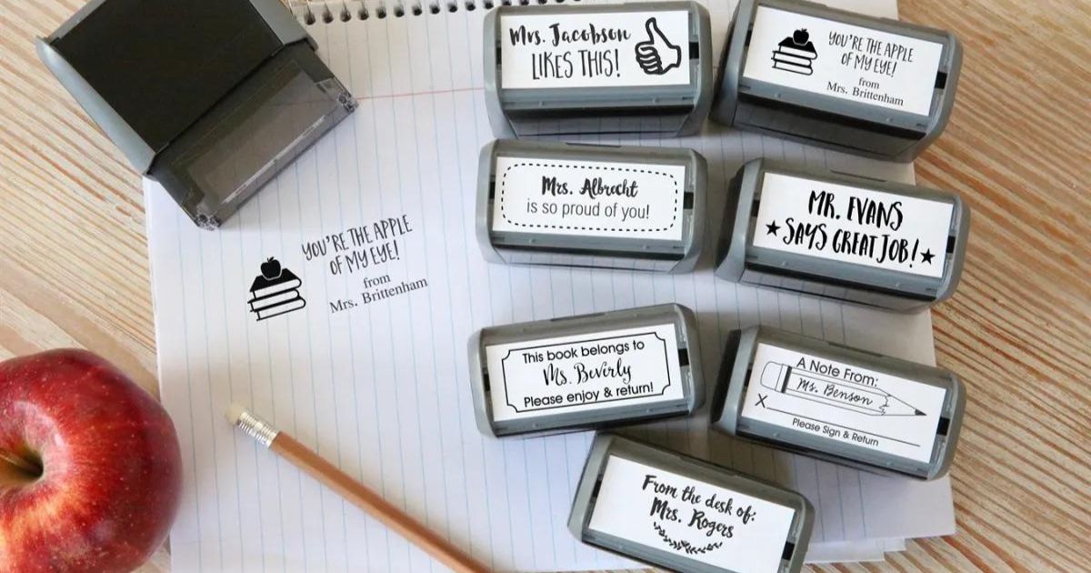 collection of self inking stamps with teacher designs next to an apple, pencil, and sheet of paper with a sample stamp
