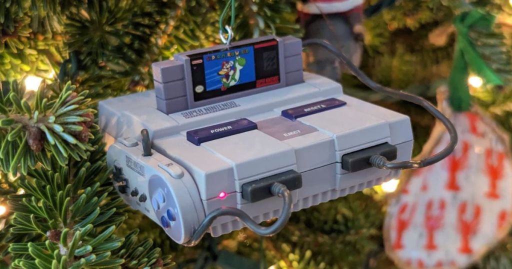 A super nintendo hallmark ornament hanging on a christmas tree with the Mario World video game inserted in the top