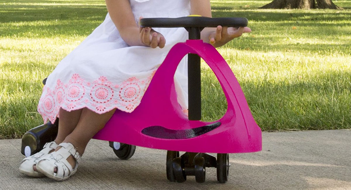 little girl wearing a dress and sandals sitting on a pink Wiggle Car Ride On Toy outside on the sidewalk near grass