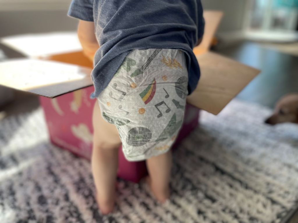 Baby standing next to a box wearing a diaper with rockstar prints on it