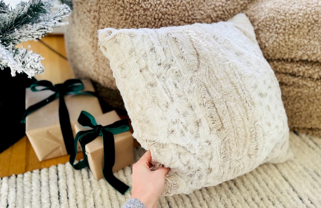 hand holding faux fur throw pillow on rug next to christmas gifts