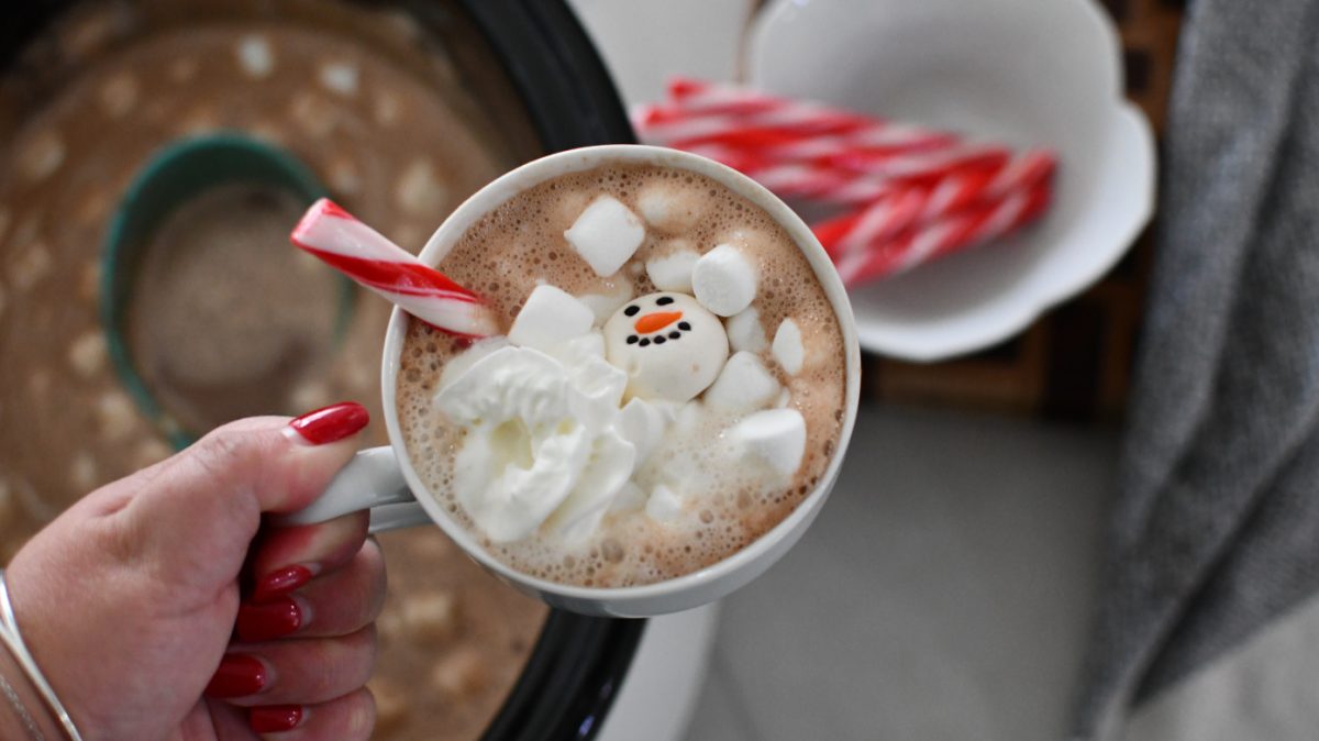 Hand holding a mug of Crock Pot Hot Chocolate with the slow cooker in background 