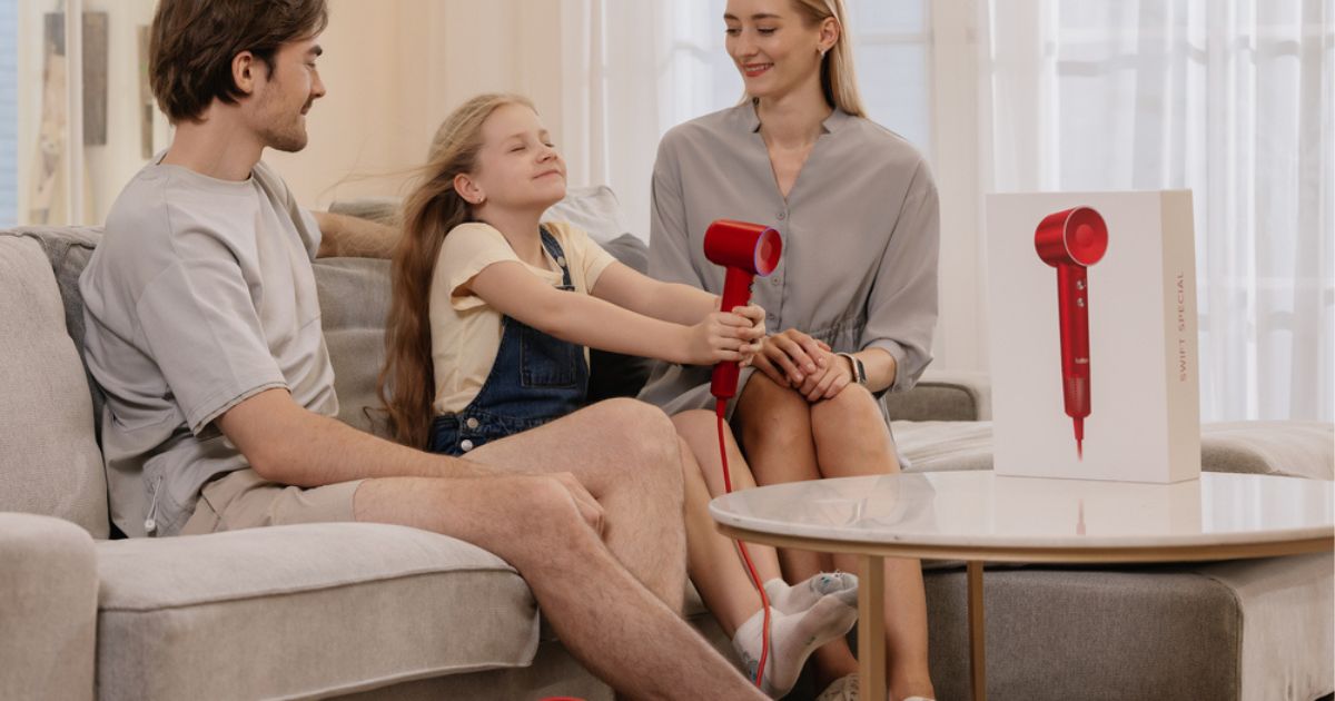 man girl and woman sitting on couch with girl using red blowdryer 