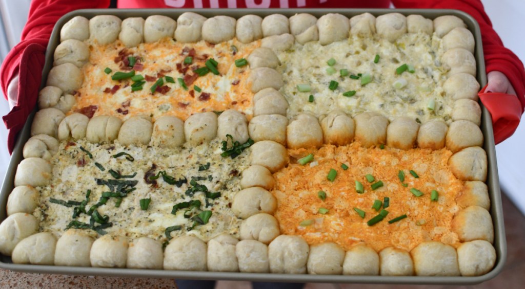 a person holding a sheet pan that contains 4 dip recipes