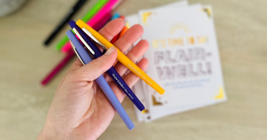 hand holding a light purple flair pen over pile of papermate pens and card