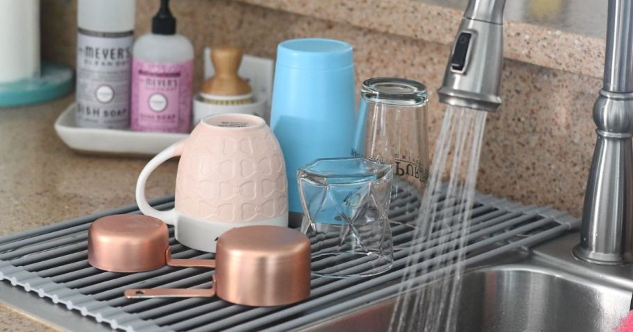 Roll up dish drying rack set on left side of a double basin sink with various glasses and measuring cups on it.