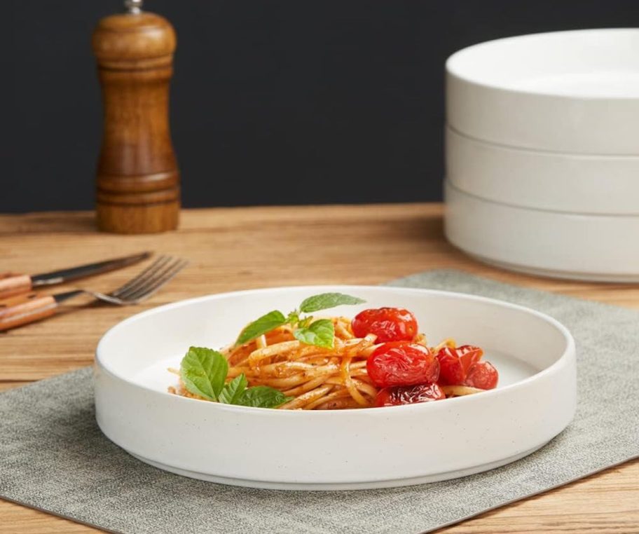 white deep pasta bowl with pasta and tomatoes in center with stack of bowl in background