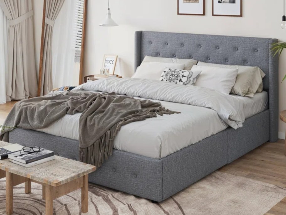 grey upholstered bed frame and headboard set with white bedding and a dark green blanket on it in a room