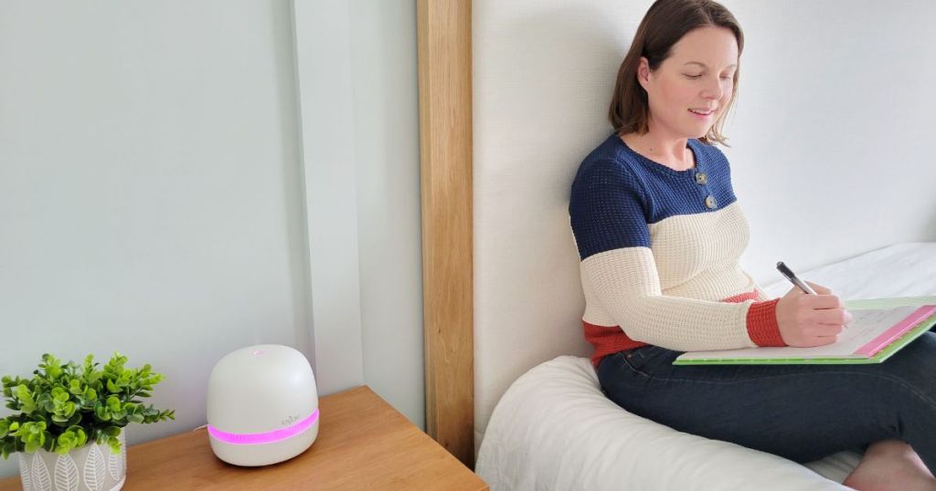 white Anjou diffuser with pink light placed on wood table beside woman laying on bed writing in notepad