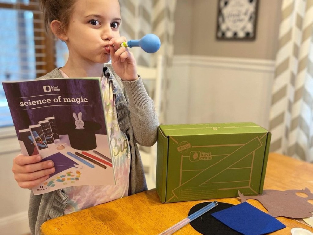 girl blowing balloon next to kiwico box