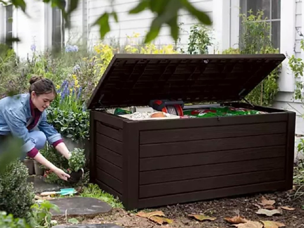 woman planting flowers next to an open Keter 165-Gallon Resin Outdoor Deck Box