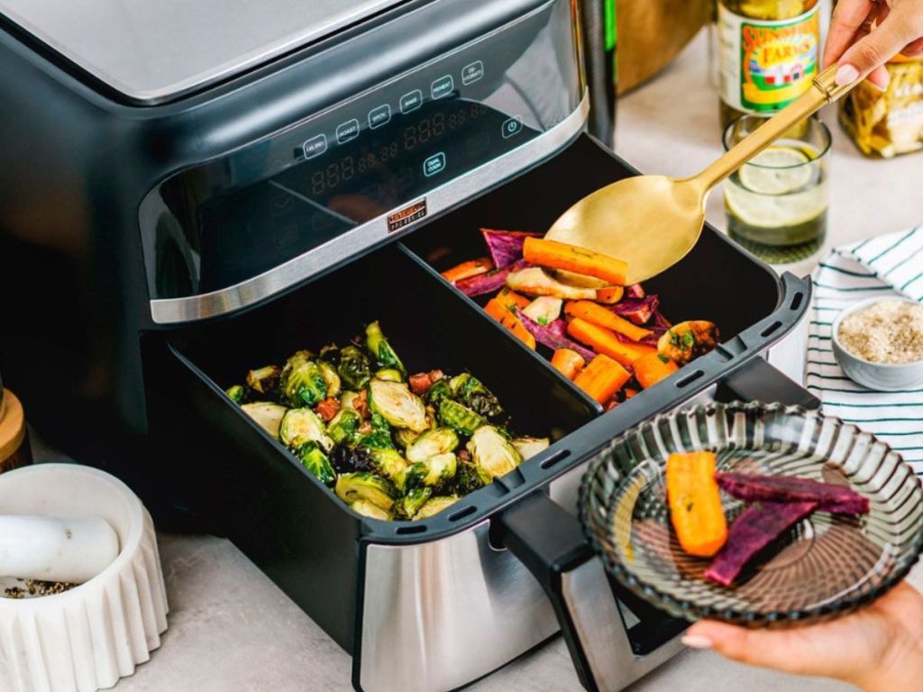 Bella Pro Series 9qt Dual Drawer Air Fryer with two kinds of veggies in the drawer and a hand scooping some carrots from the right side onto a plate with a spoon