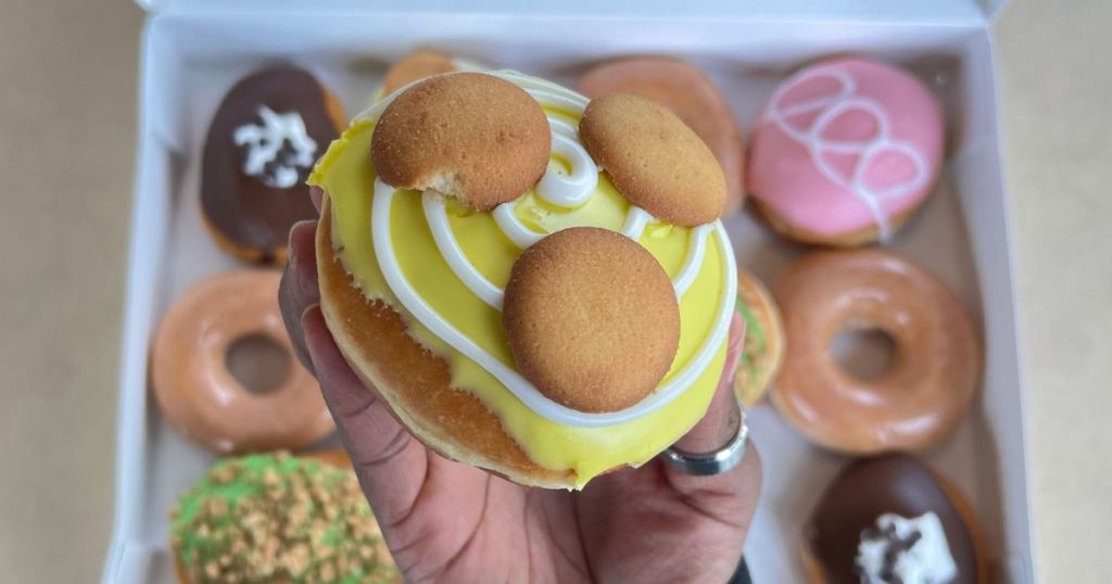 Hand holding a Banana Kreme Pie Doughnut from Krispy Kreme over a box of their limited edition fan favorite doughnuts