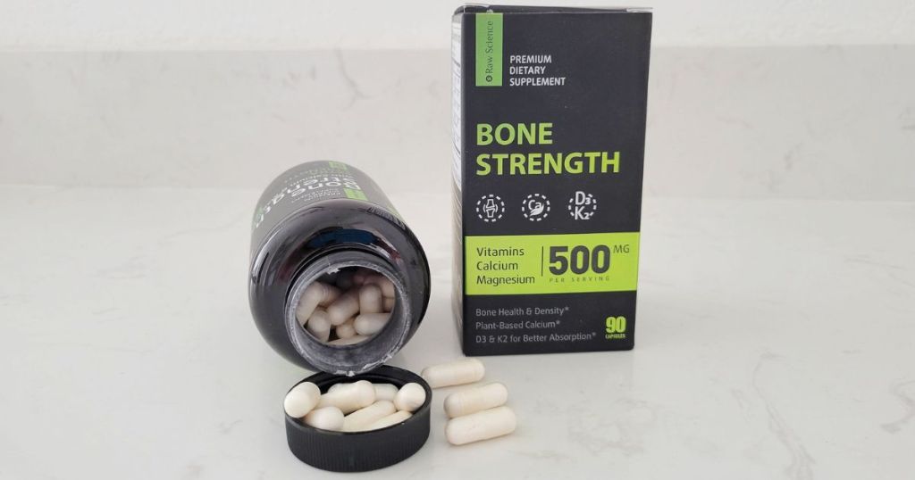 Bottle of Raw Science Bone Strength supplement tipped over with capsules in the lid, 3 capsules next to the jar with the box they come in set next to them. 