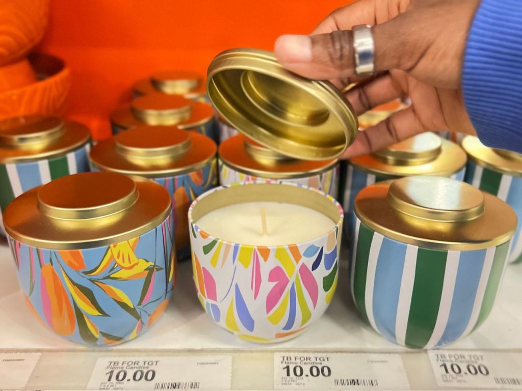 Three different Tabitha Brown Scented Candle Tins with a hand lifting the lid on one to expose the candle inside