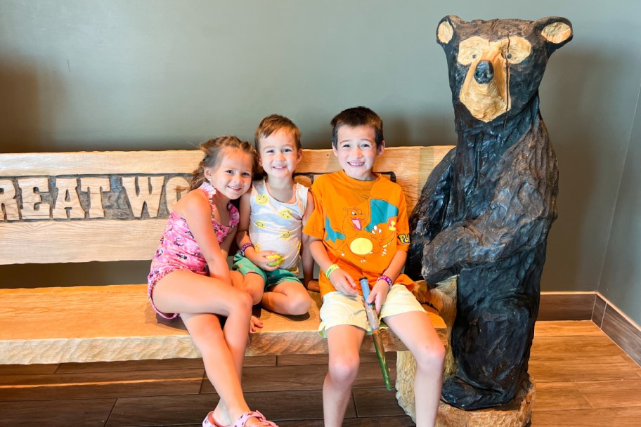 three kids sitting on bench next to wooden bear statue