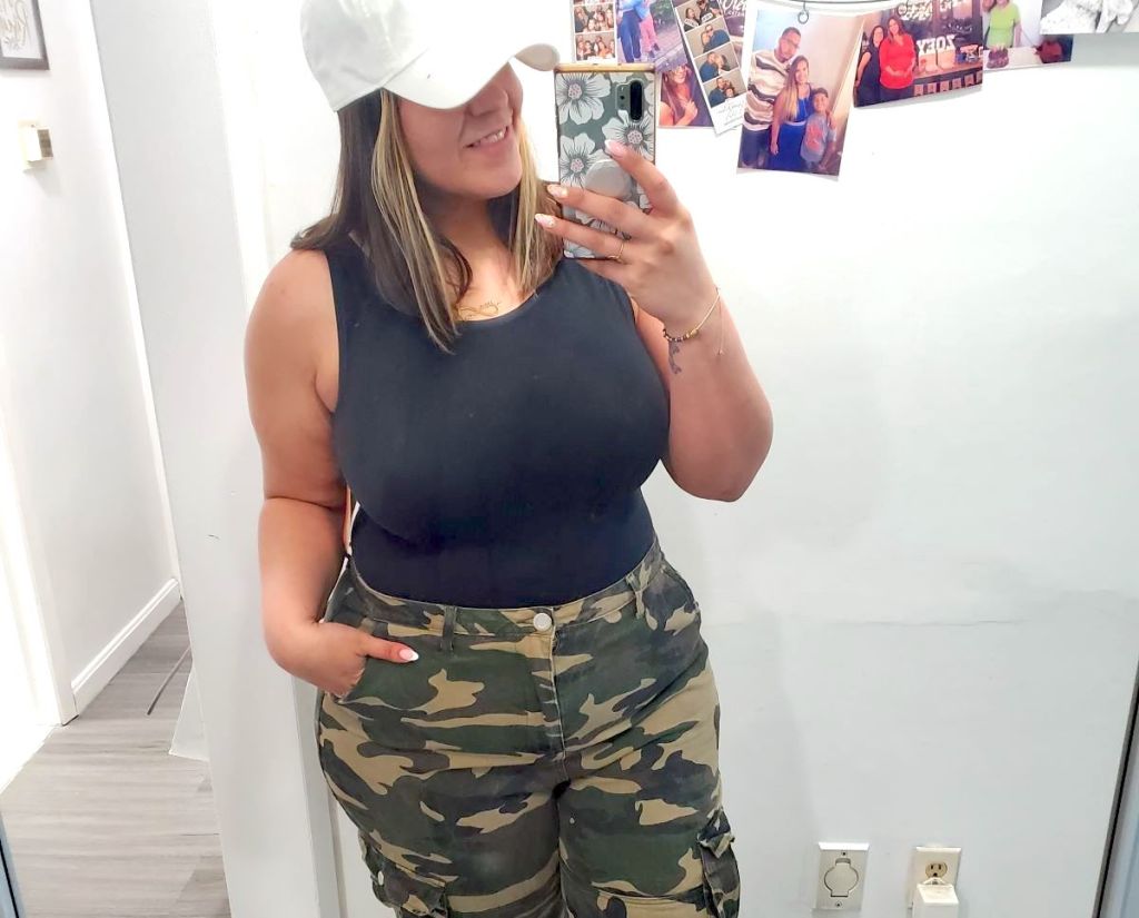 woman taking mirror selfie wearing black bodysuit hat and camo pants