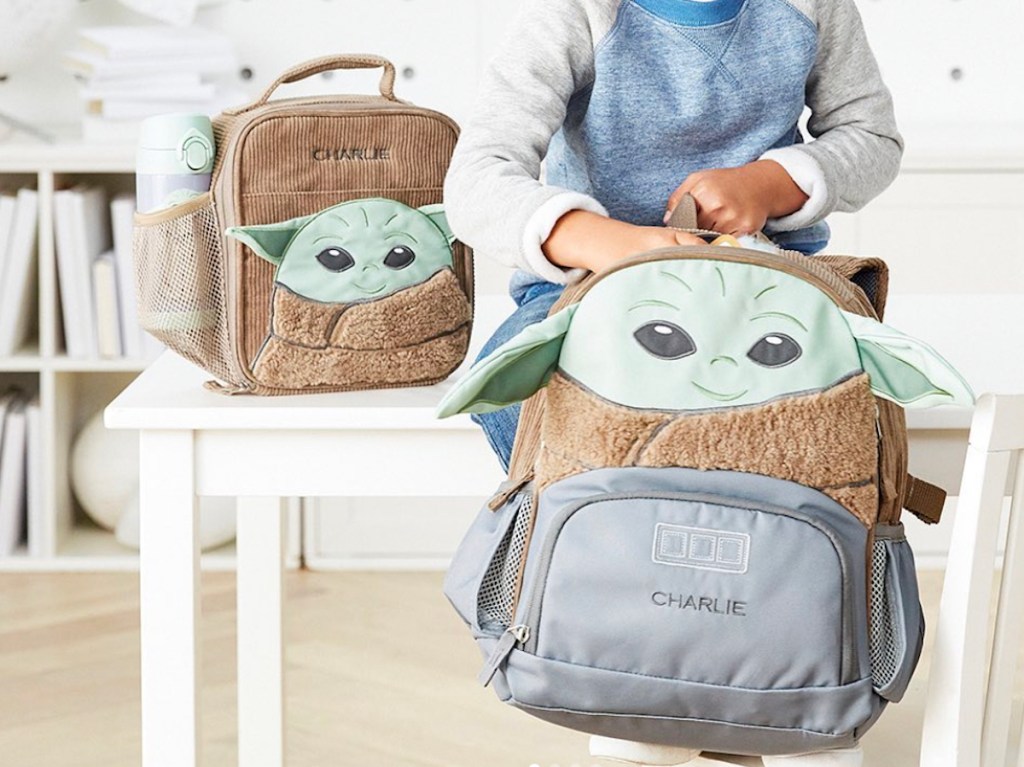 boy sitting on white kids table with brown and gray star wars lunch box and backpack