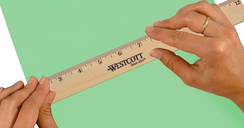 a womans hands holding a westcott 12 inch wooden ruler to measure a green piece of paper