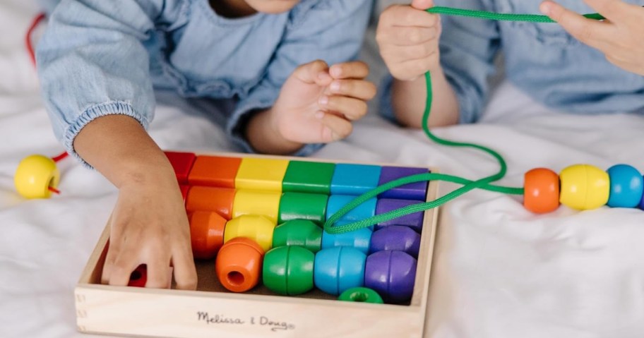 two kids laying on a bed playing with a set of wooden primary color beads and green lacing strings in a wooden box that says "Melissa & Doug"