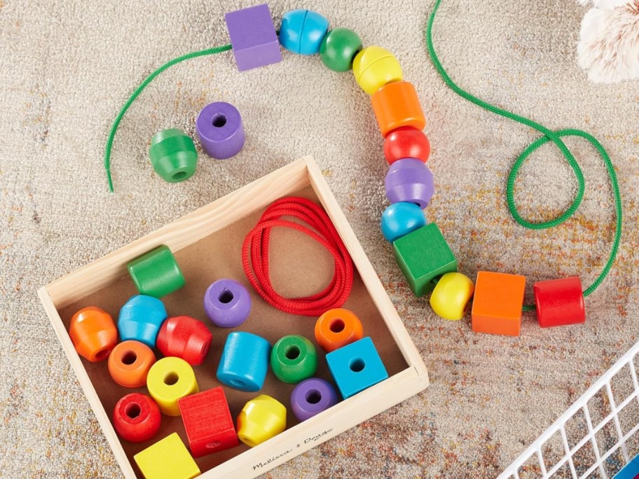 a set of wooden primary color beads in a variety of shapes in a wood tray next to a green lace string with some of the beads on it and around it