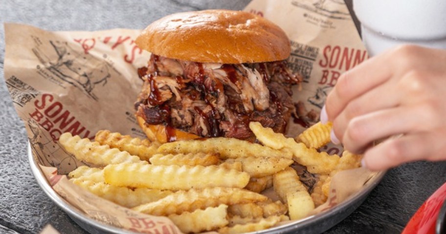 hand grabbing fry from plate with bbq sandwich and fries