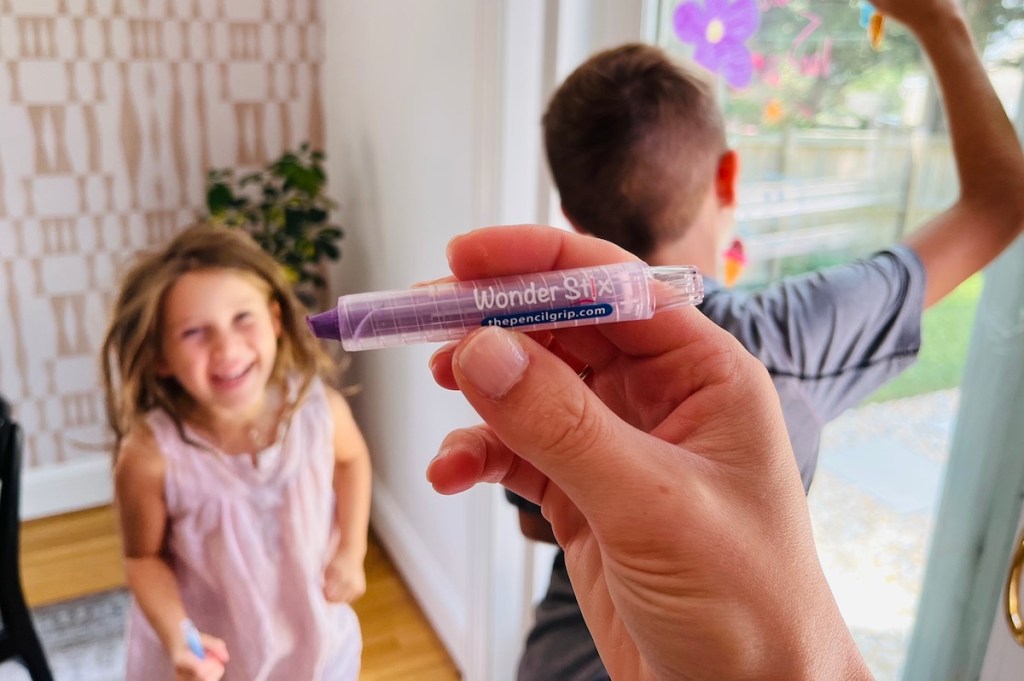 Hand holding a wonder, stix crayon in front of kids drawing on window