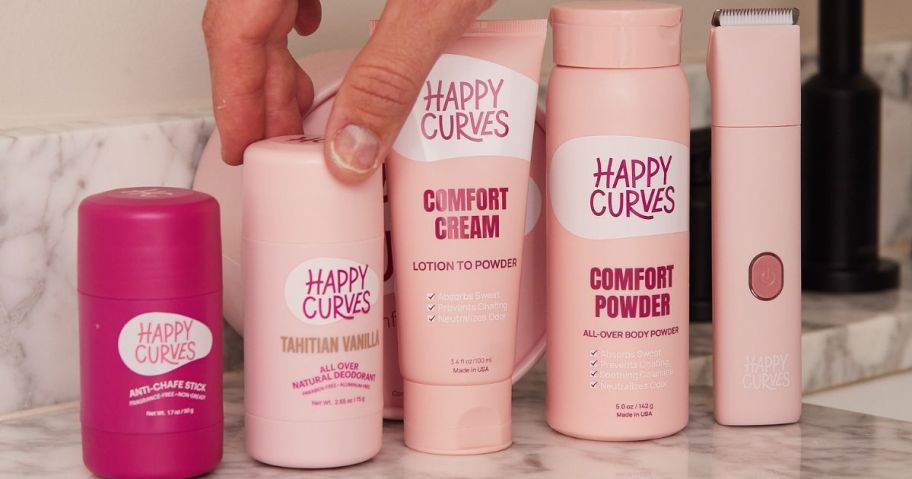 Happy Curves products lined up on a bathroom counter with a hand grabbing the deodorant stick