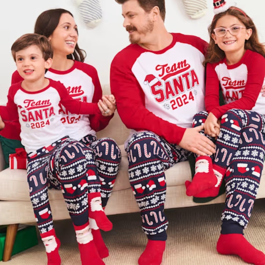 family sitting on a couch wearing matching blue, red and white Christmas pajamas that say "Team Santa"