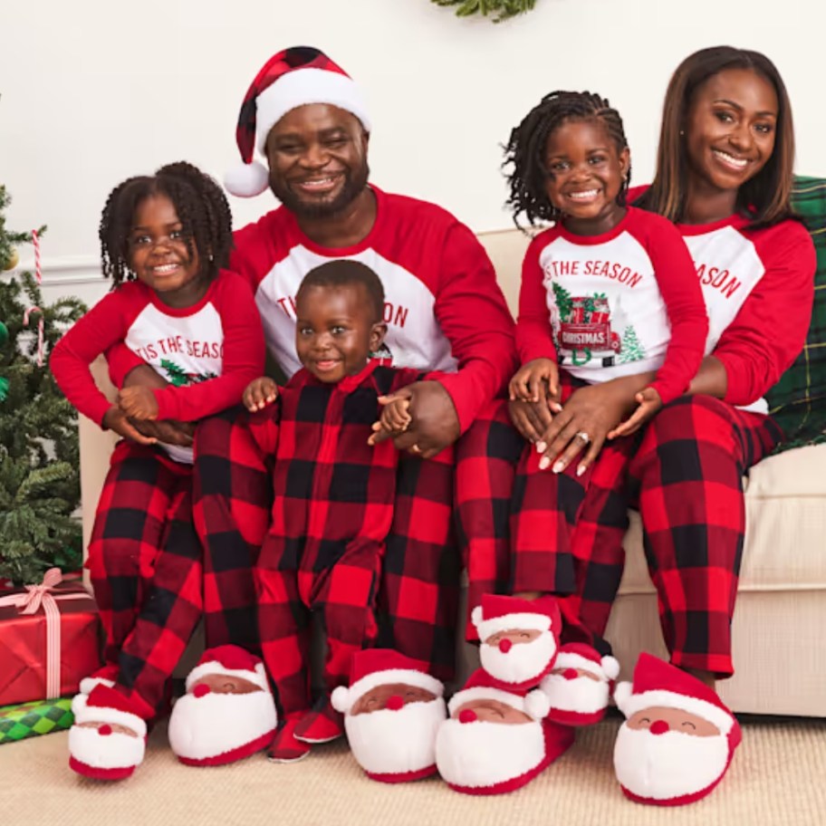 family with mom, dad, 2 little girls and baby boy wearing matching red and black plaid Christmas pajamas that say "Tis the Season"