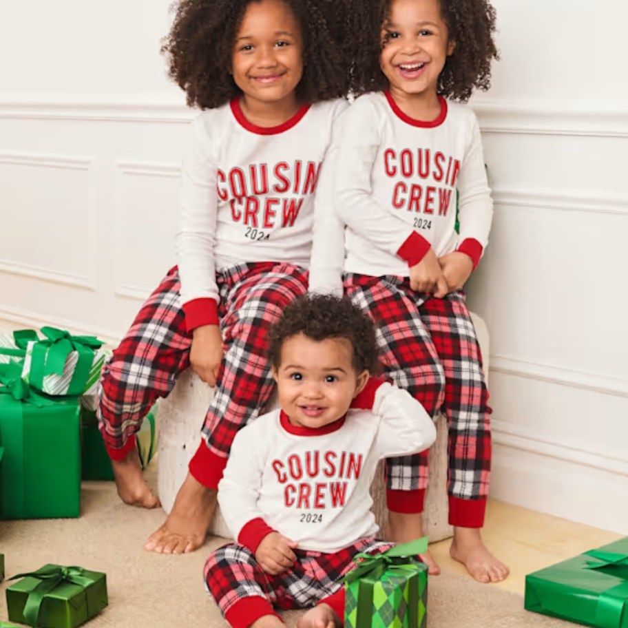 a baby and 2 little girls wearing matching red plaid and white long sleeve pajamas that say "Cousin Crew"