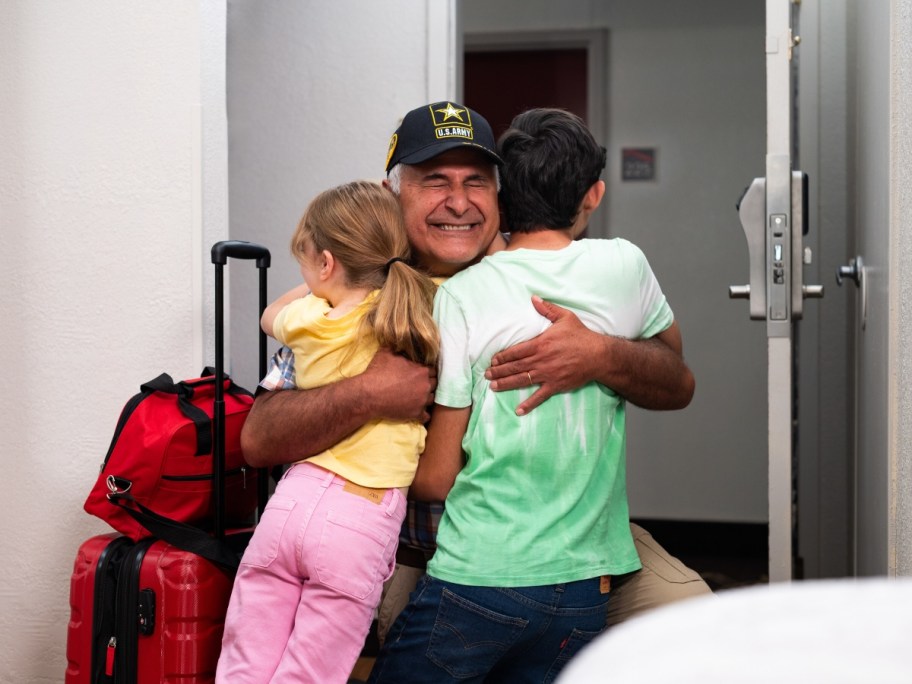 man in hat hugging children next to red luggage