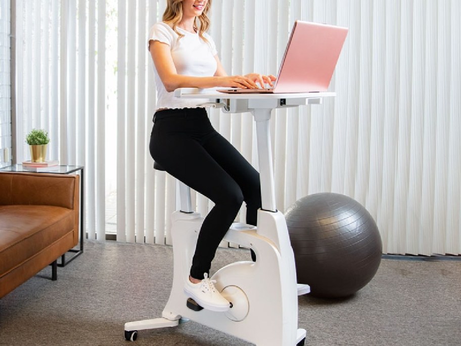woman sitting on exercise bike in white working on her laptop