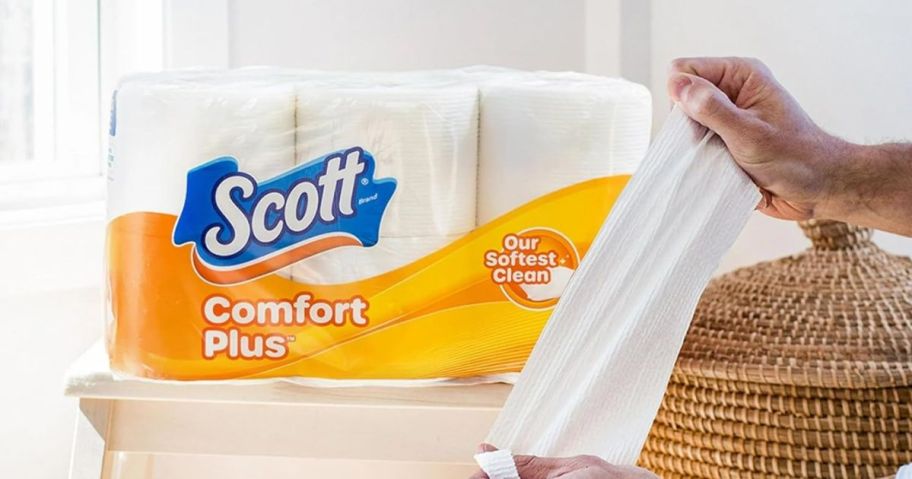 A person holding some toilet paper with a package of toilet paper in the background