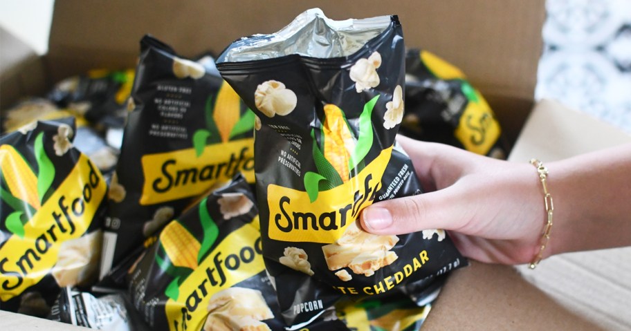 hand holding a bag of smartfood popcorn above a box full of more bags