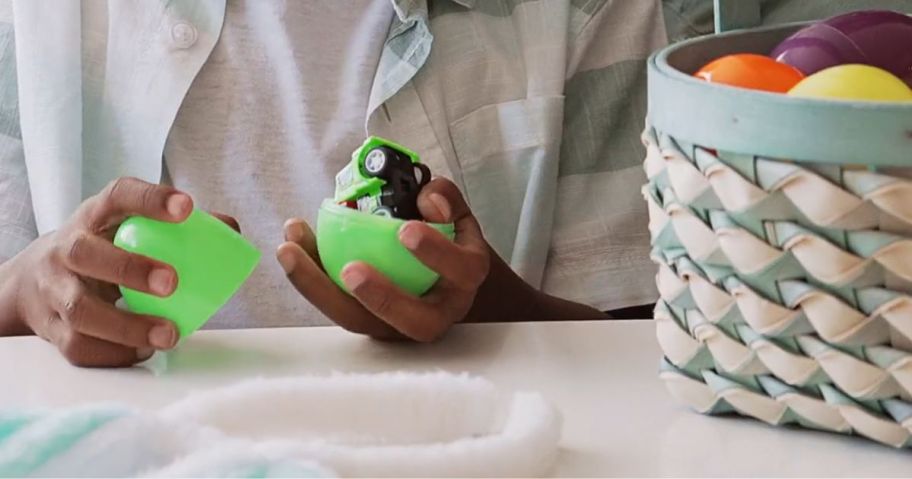 little boy's hands opening a green plastic easter egg with a toy vehicle inside, Easter basket with more eggs beside him