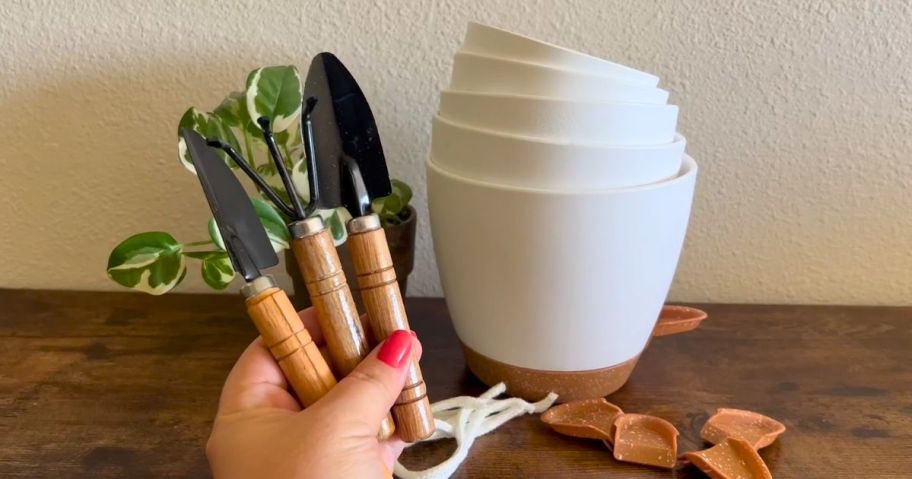 hand holding small garden tools with white self watering flower pot planters stacked in the background