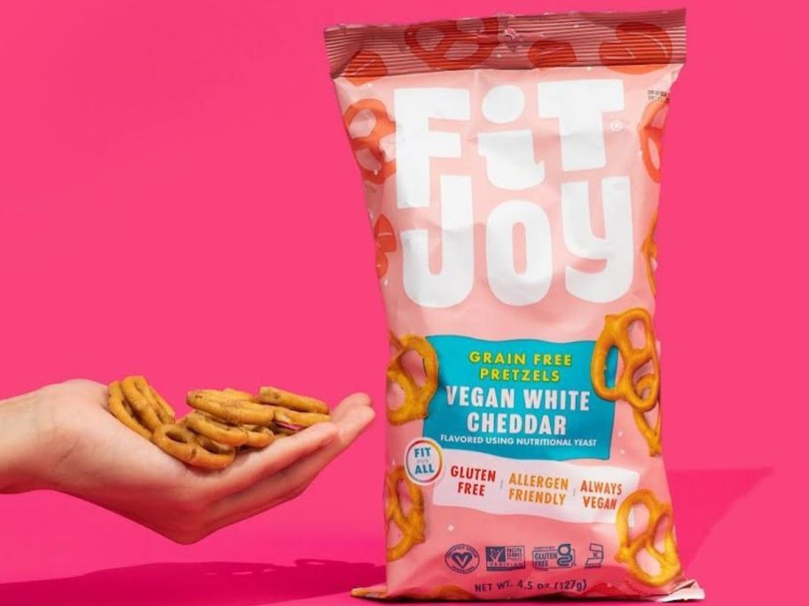 Hand holding Fit Joy White Cheddar Pretzel Twists next to a full bag