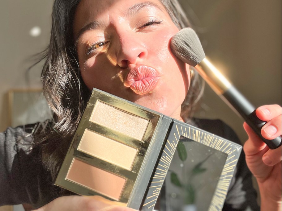 woman holding up highlighter palette and brush to her face