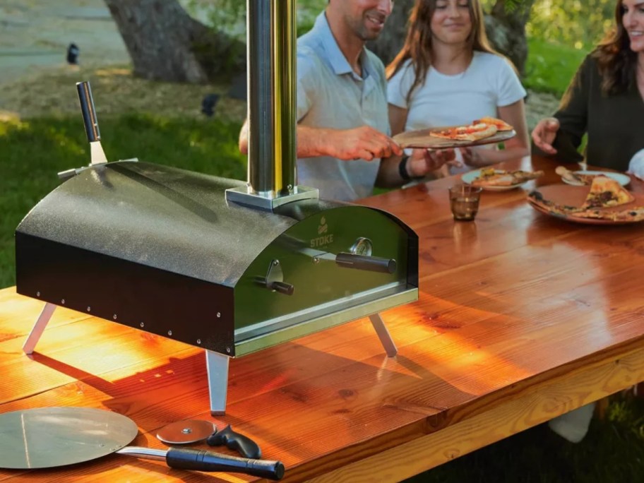 black and steel wood pizza oven sitting on outdoor table with people in the background eating pizza