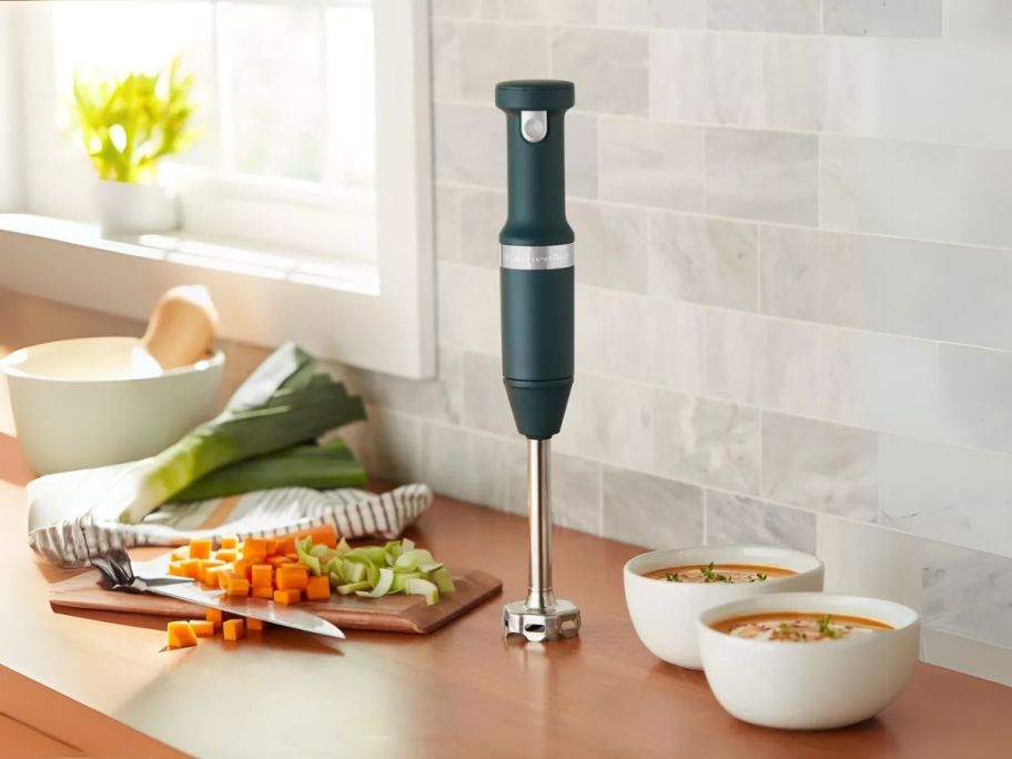 green kitchenaid cordless hand blender on counter with vegetables and bowls of soup next to it