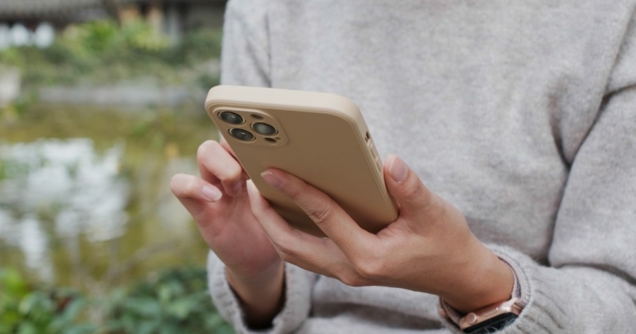 woman wearing a grey sweater sitting outside texting on a phone with a tan color case