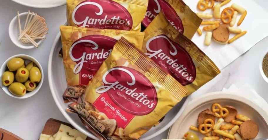 a bowl with individual Gardetto's Snack Mix bags in it on a table with some of the snack mix, bowls of olives and other foods around it