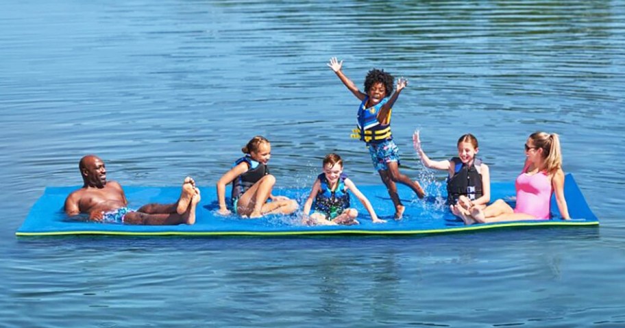 family lounging and playing on large blue floating pad in middle of lake