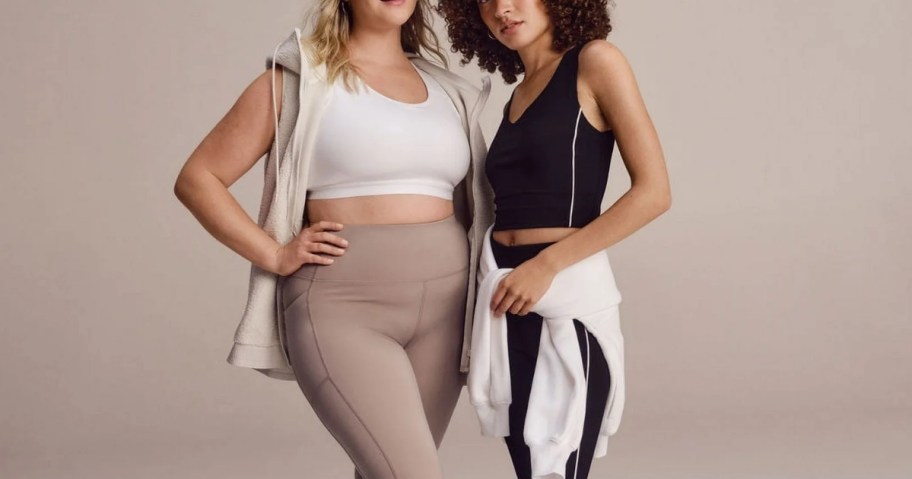 woman wearing a white sports bra and tan leggings with a sleeveless jacket and woman woman wearing a black sports bra and leggings with a white sweatshirt around her waiste