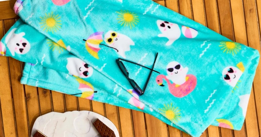 a green throw blanket with ghosts in flamingo floats and with umbrellas and cocktails on it, laying on a wood deck