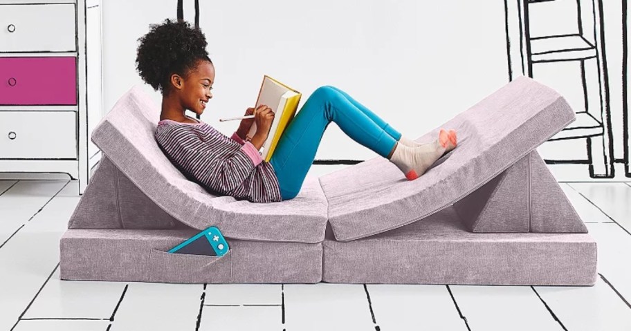 little girl reading a book sitting on a light pink folding couch with separate rectangle and triangle cushions